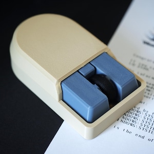 May include: A vintage computer mouse in a cream color with blue accents. The mouse has a rectangular shape with a rounded top and a black roller. The text "WINDOWS SYSTEM" is visible on a paper in the background.