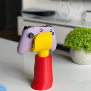 May include: A colourful controller stand in red, yellow, and purple, holding a purple Xbox controller. The stand has a red base, a yellow support, and a purple top. The controller is placed on the stand, ready for use.