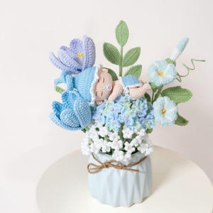 May include: A handcrafted crochet floral arrangement featuring a baby nestled among blue and white flowers. The arrangement includes blue tulips, hydrangea, and other floral elements in a light blue vase tied with twine. The baby is wearing a blue bonnet.