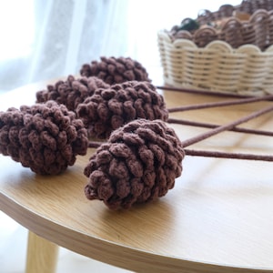 Puede incluir: Una colección de piñas de ganchillo marrón oscuro hechas a mano sobre tallos largos, dispuestas sobre una mesa de madera clara. Una cesta tejida llena de artículos similares se encuentra en el fondo, lo que sugiere un tema artesanal inspirado en la naturaleza.