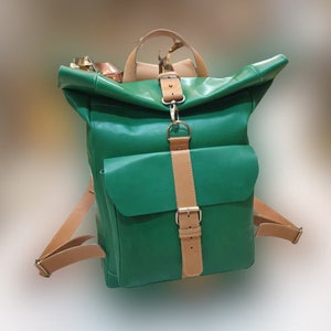 May include: A green backpack with tan leather detailing. The bag has a roll-top closure, a front pocket, and adjustable shoulder straps. It features a top handle and a buckle fastening.