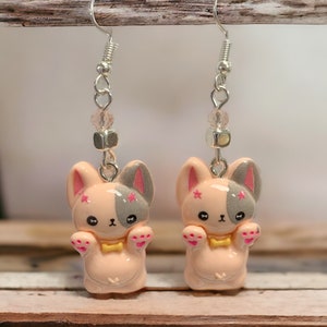 May include: A pair of dangle earrings featuring a pink and gray cartoon dog charm with pink stars and a yellow bow tie. The earrings are made with silver hooks and clear crystal beads.