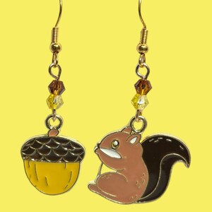 May include: A pair of gold earrings with a brown and yellow acorn charm on one earring and a brown and pink squirrel charm on the other earring. The earrings have gold beads and crystals.