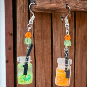 May include: A pair of dangle earrings featuring miniature glass jars filled with a limeade and an orange juice drink. Each jar has a black straw and is topped with green and orange beads.