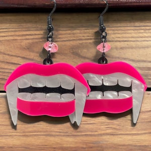 May include: A pair of pink and white acrylic earrings shaped like a vampire's mouth with fangs. The earrings have black details and a pink bead on the top of the earring hook.