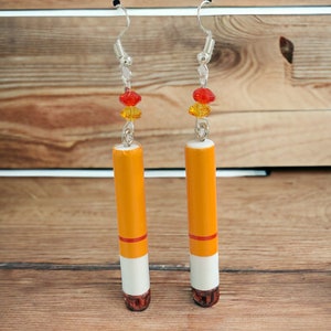 Op de afbeelding: Een paar hangende oorbellen met een sigaretvormige hanger in een kleurenschema van rood, wit en geel. De oorbellen zijn gemaakt van zilverkleurig metaal en hebben een rood, geel en oranje kralenaccent.
