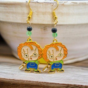 May include: Gold-tone earrings featuring a cartoon character with red hair, a blue shirt, and a knife. The earrings have green and black beads.