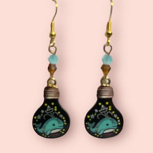 May include: A pair of gold dangle earrings with a black lightbulb charm featuring a blue whale and a small sailboat. The lightbulb is filled with gold glitter and stars.