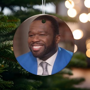 May include: Circular ceramic Christmas decoration featuring a smiling man in a blue suit and grey tie. The ornament has a white hanging string and is set against a blurred background of Christmas tree branches and lights.