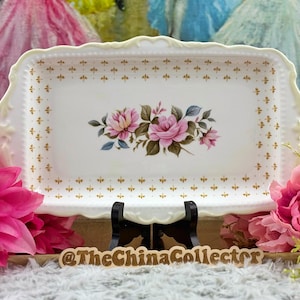 May include: A rectangular white porcelain serving tray featuring a central floral motif of pink roses with green leaves, surrounded by a delicate gold fleur-de-lis border pattern. The tray has scalloped edges and a cream-colored rim.
