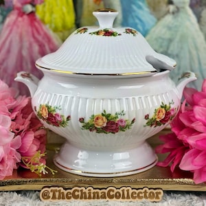 Puede incluir: Una sopera de porcelana blanca con tapa, adornada con un motivo floral de rosas rojas, amarillas y rosas. La sopera tiene ribetes y asas doradas. La base es redonda y elevada. El fondo incluye flores rosas.