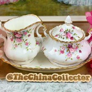 Può includere: Un set di crema e zuccheriera in porcellana bianca con un motivo floreale a rose e bordi dorati. Il set comprende una caraffa per la panna, una zuccheriera con coperchio e un vassoio abbinato. Il motivo floreale presenta rose rosa e rosse con foglie verdi.