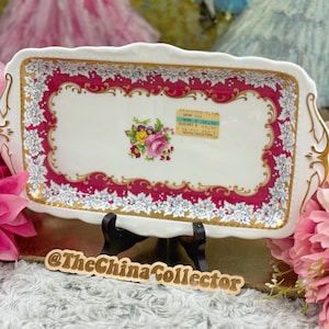 May include: A rectangular, white ceramic serving tray with a floral design. The tray features a red border with white floral accents and gold trim. A cluster of flowers is in the center. The tray has handles and is on a black stand.