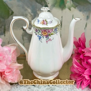 Puede incluir: Una cafetera de porcelana blanca con un diseño floral y ribetes dorados. La cafetera tiene un asa curvada y un pico largo. La tapa está rematada con un pequeño diseño floral. Flores rosas en primer plano.