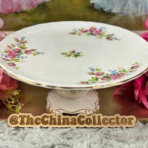 May include: A white porcelain cake stand with a scalloped edge and gold trim. Decorated with pink roses, blue flowers, and green leaves. The base is white with a decorative design.