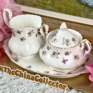 May include: A white porcelain tea set with a creamer, lidded sugar bowl, and serving tray. The set is decorated with purple floral patterns and gold trim. The tray measures approximately 10 inches in length.