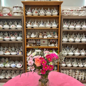 Op de afbeelding: Een verzameling vintage theepotten en serveerschalen tentoongesteld op houten planken. De theepotten hebben bloemmotieven en ingewikkelde patronen. Een boeket roze en oranje bloemen in een glazen vaas staat op een roze tafelkleed.