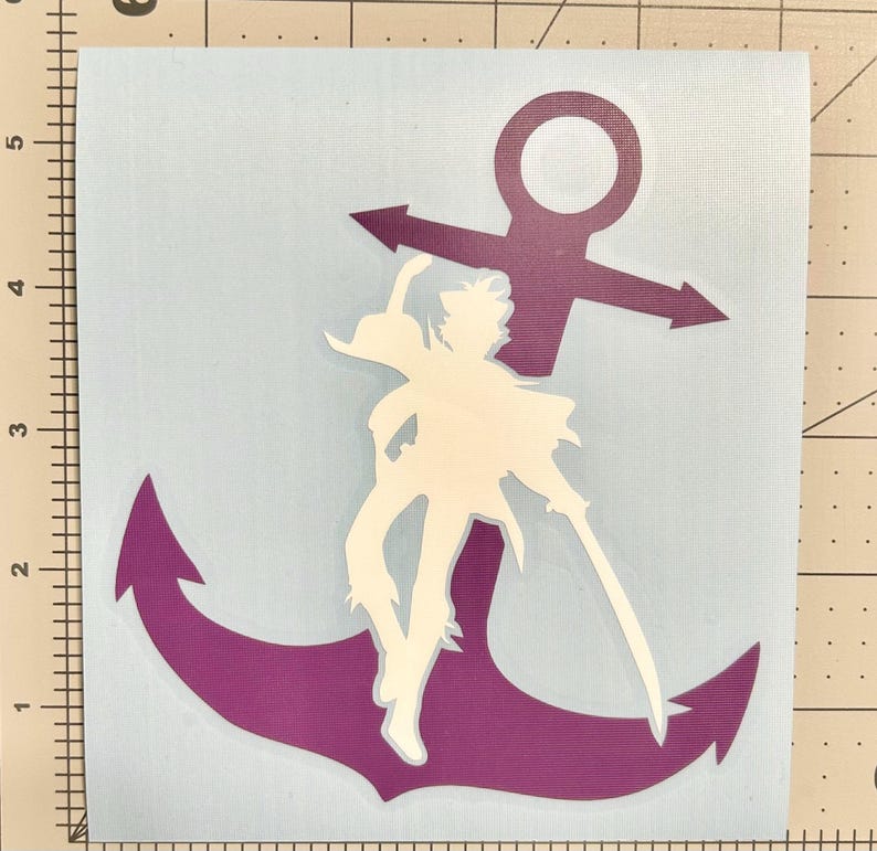 Puede incluir: Un adhesivo de vinilo morado con forma de ancla que presenta la silueta blanca de una figura pirata sosteniendo una espada, sobre un soporte azul claro con una regla visible que muestra medidas en cent&iacute;metros en los bordes izquierdo y superior.