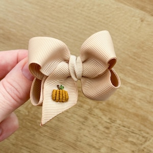 May include: A beige hair bow made of ribbed grosgrain ribbon featuring a small, embroidered orange pumpkin with a green stem on one of the bottom tails. The bow has a classic multi-loop structure with a central knot and two hanging ribbon tails.