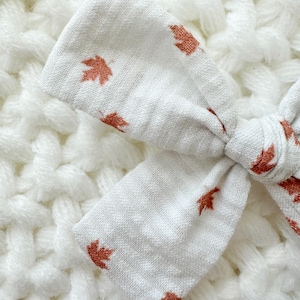May include: A white fabric bow with a repeating pattern of small, rust-colored maple leaves. The bow is knotted in the center and sits on a textured white surface, creating a soft, autumnal aesthetic. Ideal for hair accessories.