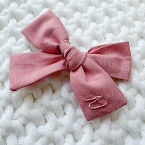 May include: A dusty rose-colored hair bow with a knotted center and two loops. The bow is made of fabric and has a stitched letter "N" on one of the loops. The bow is lying on a white, textured surface.