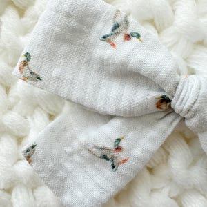 May include: A light grey fabric bow featuring a repeating pattern of mallard ducks. The ducks have green, brown, and red details. The bow is knotted and rests on a white, textured background.