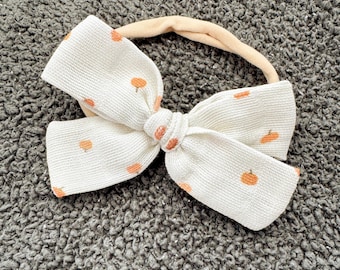 May include: A cream-coloured fabric headband featuring a large, structured bow. The material has a subtle ribbed texture and is decorated with a repeating pattern of small, orange pumpkin icons scattered across the surface of the bow and band.