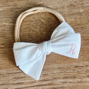 May include: White fabric bow headband with a pink embroidered letter "R" on the bow.