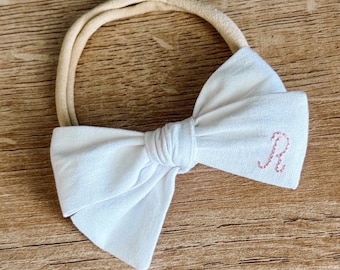 Op de afbeelding: Een wit stoffen strik hoofdband met een roze geborduurde letter "R" op de strik.