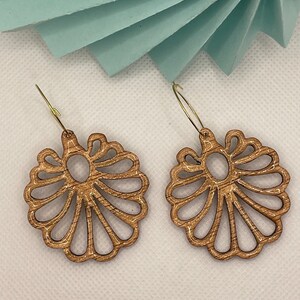 Puede incluir: Un par de pendientes de madera con un diseño delicado e intrincado. Los pendientes tienen forma de flor con un centro circular y múltiples capas de pétalos. La madera es de color marrón claro y los pendientes tienen ganchos de color dorado.
