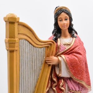 May include: A figurine of a woman playing a harp. She is wearing a white dress with a red and pink robe. The harp is gold and has a white stringed section. The figurine is standing on a black base.