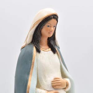 May include: A statue of a woman in a white dress and a blue cloak, with a white head covering. The woman is holding her hands over her abdomen. The statue has gold accents and dark hair.