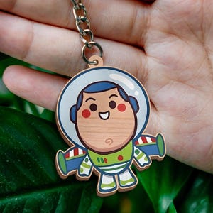 May include: A wooden keychain featuring a cartoon space ranger character with a white helmet, blue hood, green chest plate, and purple wings. The character has a light wood grain face, red cheeks, and a smiling expression, attached to a metal ring.