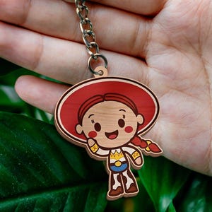 May include: A wooden keychain featuring a cartoon character with red hair in braids, a large red hat, a white shirt with yellow and blue details, and brown cow-print pants. The character is attached to a silver metal split ring by a short chain.