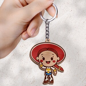 May include: A wooden keychain featuring a cartoon character with red hair in a braid, wearing a large red hat, a white shirt with yellow accents, a blue belt, and cow-print pants. The character has a smiling expression and is attached to a metal ring.