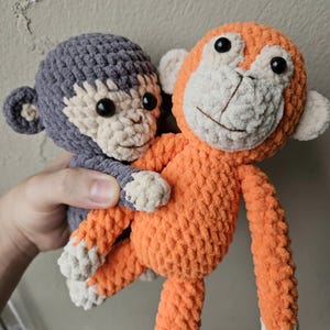 以下が含まれることがあります： 手作りの2匹のかぎ針編みの猿。1匹はグレー、もう1匹はオレンジ色です。どちらも白い顔、耳、足、黒いボタンの目があります。オレンジ色の猿は灰色の猿を抱きしめています。おもちゃは柔らかく、抱きしめたくなります。
