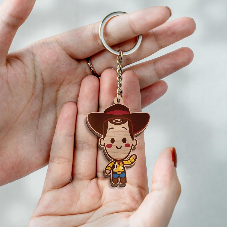 May include: A wooden keychain featuring a cartoon character with a brown cowboy hat, a yellow shirt with a cow print vest, and blue pants. The character has a light wood grain texture, a simple smiling face, and is attached to a silver metal ring.