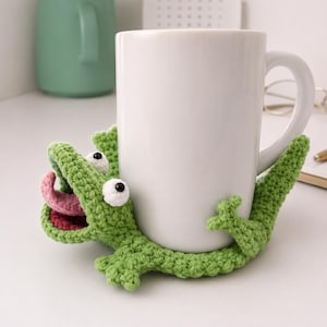 Könnte beinhalten: Eine weiße Keramiktasse wird von einem handgefertigten grünen Krokodil aus Häkelarbeit gehalten. Das Krokodil hat weiße und schwarze Augen, eine rosa Zunge und einen roten Mund. Die Tasse steht auf einer weißen Oberfläche, mit einer grünen Tasse im Hintergrund.