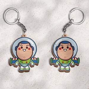 May include: Two identical wooden keychains featuring a cartoon astronaut character with a light wood grain face, a blue helmet, and a green, white, and blue space suit with red accents. Each keychain includes a silver metal ring and chain.