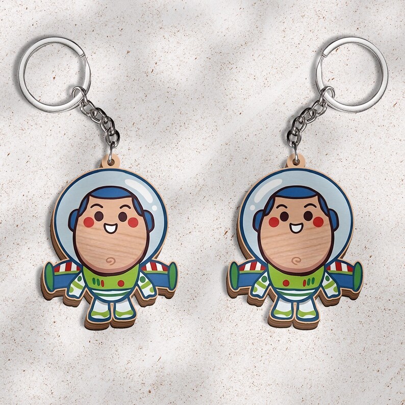 May include: Two identical wooden keychains featuring a cartoon astronaut character with a light wood grain face, a blue helmet, and a green, white, and blue space suit with red accents. Each keychain includes a silver metal ring and chain.