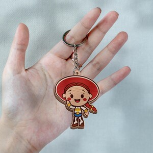 May include: A wooden keychain featuring a cartoon character with a large red hat, red braided hair, and a cow-print outfit. The character has a smiling face with rosy cheeks and is attached to a silver-colored metal split ring by a short chain.