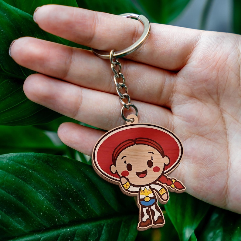 May include: A wooden keychain featuring a cartoon character with red hair in braids, a large red hat, a white shirt with yellow and blue details, and brown cow-print pants. The character is attached to a silver metal split ring by a short chain.
