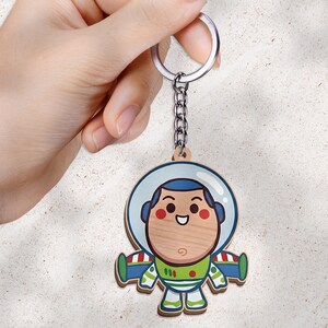 May include: A wooden keychain featuring a cartoon astronaut character with a round face, a blue hood, and a green and white space suit. The character has a smiling expression with rosy cheeks. The keychain includes a silver metal ring and chain.