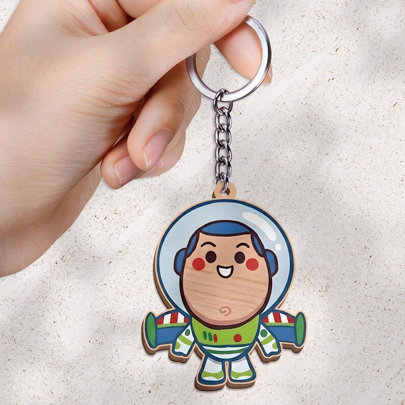 May include: A wooden keychain featuring a cartoon astronaut character with a round face, a blue hood, and a green and white space suit. The character has a smiling expression with rosy cheeks. The keychain includes a silver metal ring and chain.