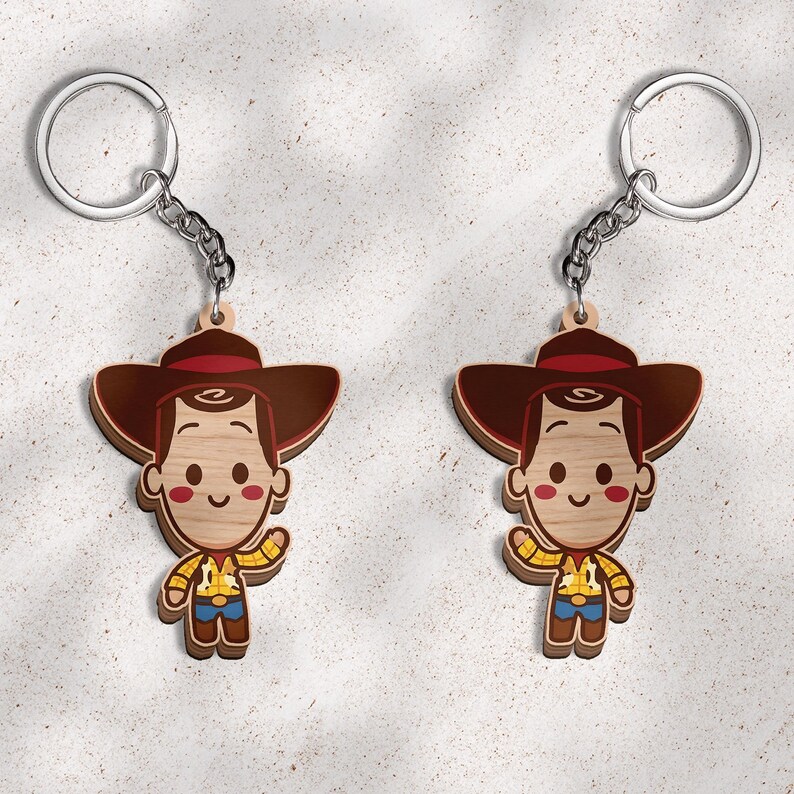 May include: Two identical wooden keychains featuring a cartoon cowboy character with a brown hat, a yellow patterned shirt, blue pants, and brown boots. Each keychain includes a silver metal split ring attached by a short silver chain.