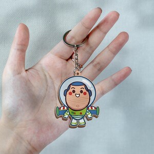 May include: A wooden keychain featuring a cartoon character with a round face, wearing a white space helmet, green and purple wings, and a white suit with red and green accents. The character has a smiling expression and is attached to a metal ring.