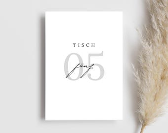 May include: A white card with the text "Tisch 05 fünf" in black and gray lettering. The card is a table number for a wedding or other event.