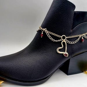 May include: Black ankle boot with a gold chain and heart charm ankle bracelet. The chain has red beads.
