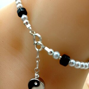 Peut inclure: Un bracelet argenté orné d'un pendentif Yin et Yang. Le bracelet est composé de petites perles rondes argentées et de perles carrées noires. Le pendentif est un symbole Yin et Yang noir et blanc avec un petit cristal.