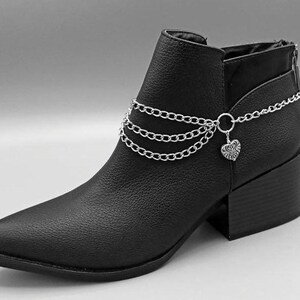 Puede incluir: Botín negro con punta y tacón cuadrado. El botín presenta una pulsera de tobillo de cadena plateada con un dije de corazón. El botín tiene una apariencia de cuero texturizado y una cremallera en la parte trasera.